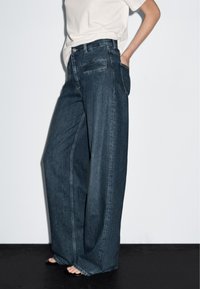 Jeans in denim scuro a gamba larga con vita alta, dotati di tasche frontali e dettagli di cucitura discreti, appoggiati su una superficie nera.