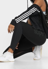 Person som bär svarta Adidas leggings, svart jacka med vita ränder och vita Adidas sneakers, hukande mot vit bakgrund.