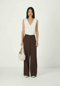 Vit ärmlös topp med V-ringning och knappar, parat med högmidjade bruna vida byxor. Modellen håller en ljusbeige väska.