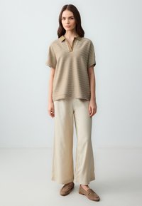 Polo shirt beige chiaro a righe orizzontali, abbinato a pantaloni a gamba larga color crema e mocassini marroni, su uno sfondo neutro.