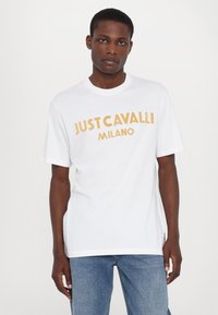 Bílý bavlněný tričko s výrazným žlutým nápisem "JUST CAVALLI MILANO", kulatým výstřihem a krátkými rukávy. Kombinováno s modrými džíny.