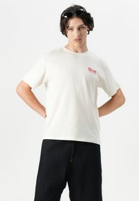 Cotton crew neck t-shirt in off-white with short sleeves. Features red text graphic on the left chest. Paired with dark denim pants.
