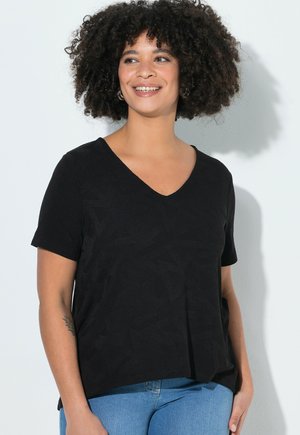 Smilende kvinde med krøllet hår iført en sort V-hals kortærmet skjorte og blå jeans, stående mod en hvid baggrund.