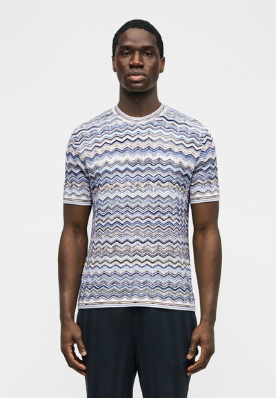 Άνδρας που φοράει κοντομάνικο πουκάμισο με πολύχρωμο ζιγκ-ζαγκ σχέδιο και σκούρο παντελόνι, στέκεται μπροστά σε απλό λευκό φόντο.