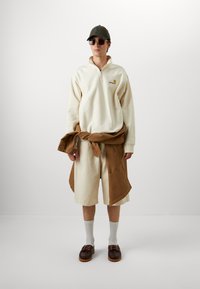 Sweatshirt zippé beige clair, shorts marron superposés, chaussures marron, chaussettes blanches, casquette de baseball verte, lunettes de soleil. Style simple et décontracté.