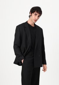 Black double-breasted blazer in a smooth fabric, featuring notched lapels and two front buttons, styled with a black t-shirt and trousers.