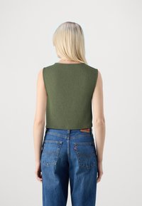 Blondyn noszący bezrękawnik w kolorze oliwkowym i wysokie niebieskie dżinsy, pokazany od tyłu na białym tle.