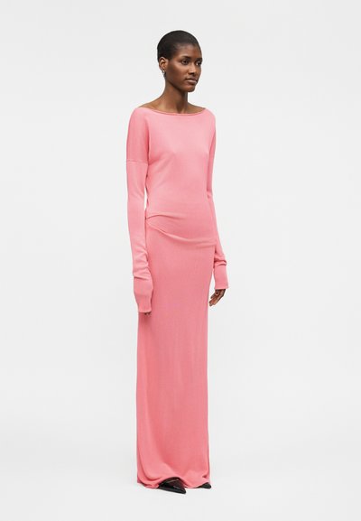 Γυναίκα όρθια, φοράει μακρυμάνικο φόρεμα μακριά ροζ και μαύρα μυτερά παπούτσια σε απλό λευκό φόντο.