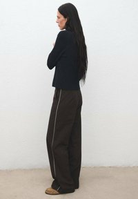 Donna con lunghi capelli scuri, che indossa un maglione nero, pantaloni larghi marrone scuro con strisce bianche ai lati e scarpe mocassino marroni, in piedi di profilo.
