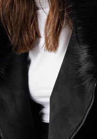 Largo cabello castaño sobre una blusa blanca, parcialmente cubierto por una chaqueta negra con un cuello de pelo texturizado y una cremallera visible.