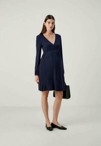 Vestido de maternidade azul marinho com mangas longas, decote em V e cintura franzida. Tecido suave, até ao joelho, combinado com uma mala preta e sapatos planos.