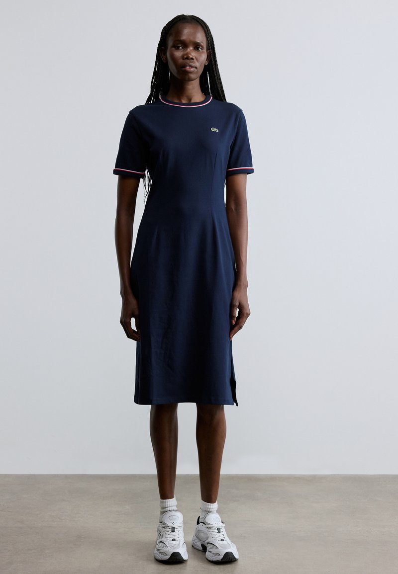 Tumši zila kleita ar īsām piedurknēm, kontrastējoša sarkana apmale pie kakla un piedurknēm; pieguļošs korsete un paplašināta apakša; kokvilnas maisījuma audums.