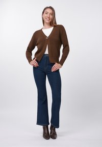 Καφέ πλεκτή ζακέτα με ρίγες, V-λαιμόκοψη και διακοσμητικό κουμπί, συνδυασμένη με ψηλόμεσα σκούρα μπλε τζιν με καμπάνα και καφέ μποτάκια αστραγάλου.