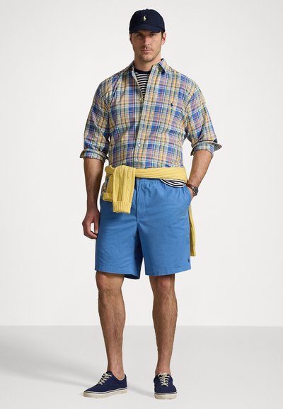 Chemise à carreaux bleu, jaune et vert ; pull jaune noué à la taille ; shorts bleus ; baskets navy ; casquette de baseball.
