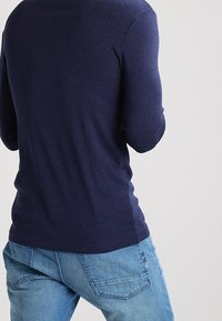 Muž oblečený v námořnickém tričku s dlouhým rukávem a světle modrých džínách, zobrazený zezadu na jednobarevném světlém pozadí.