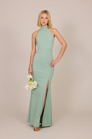 Naine varrukateta mintrohelises kõrge kaelusega kleidis reite lõhikuga, hoides valgeid kalla lillet neutraalse tausta ees.