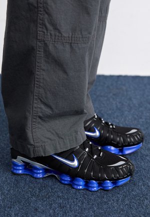 Chaussures de sport Nike noires et bleues portées avec un pantalon gris foncé sur un sol en moquette bleue.