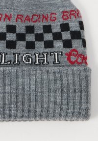 Gorro de punto gris con textura acanalada, patrón a cuadros rojo y negro, y texto en blanco. La tela parece suave y cálida.
