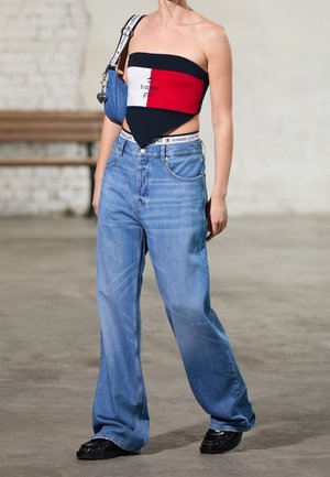 Jeune femme portant un haut bandeau Tommy Girl rouge, blanc et noir, un jean bleu ample, des chaussures noires et un sac à bandoulière en denim.