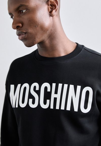 Sudadera negra con letras en blanco en negrita "MOSCHINO" en el frente. Textura suave con cuello redondo y mangas montadas.