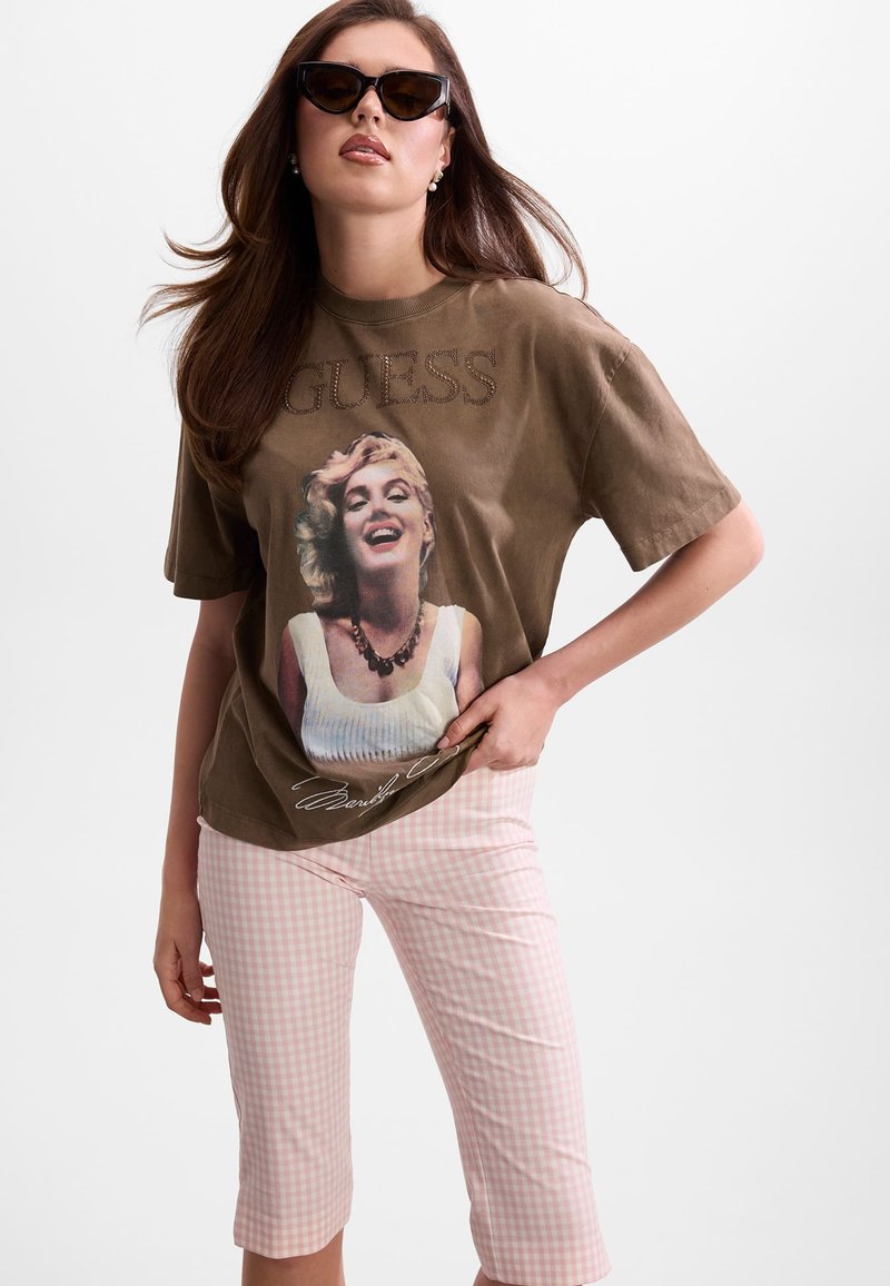 Femme portant un t-shirt marron oversize avec un imprimé Marilyn Monroe et le texte "GUESS", un pantalon capri à carreaux rose et blanc, et des lunettes de soleil noires.