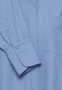 Camicia azzurra chiara a maniche lunghe, con polsini abbottonati, dettaglio arricciato sulle maniche e taschino sul petto. Tessuto dalla texture liscia.