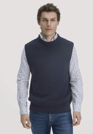 Marineblaue, gestrickte ärmellose Sweater mit hohem Kragen, getragen über einem weißen Hemd mit blauem Blumenmuster, kombiniert mit Jeans.