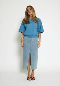 Felpa azzurra oversize con maniche corte, abbinata a una gonna di jeans azzurra chiara con spacco frontale. Il modello indossa sandali neri.