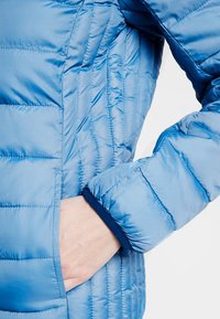 Jasnoniebieska kurtka ocieplana z pikowanym wzorem, stojącym kołnierzem i granatowymi mankietami. Posiada kieszeń po lewej stronie.