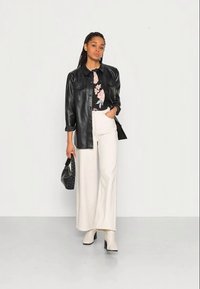 Camisa jaqueta de imitação de couro preta sobre uma blusa com padrão floral, combinada com calças brancas de perna larga e botas de cano curto bege. Acessório: bolsa preta.