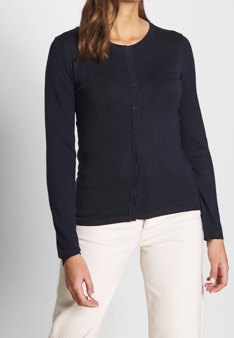 Kvinna som bär en figursydd marinblå knappad cardigan med långa ärmar och off-white byxor, visad från axlarna till höfterna.