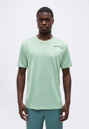 Ung mand med mørk hud iført en lysegrøn Nike træningstrøje og blågrønne shorts, stående mod en ensfarvet hvid baggrund.