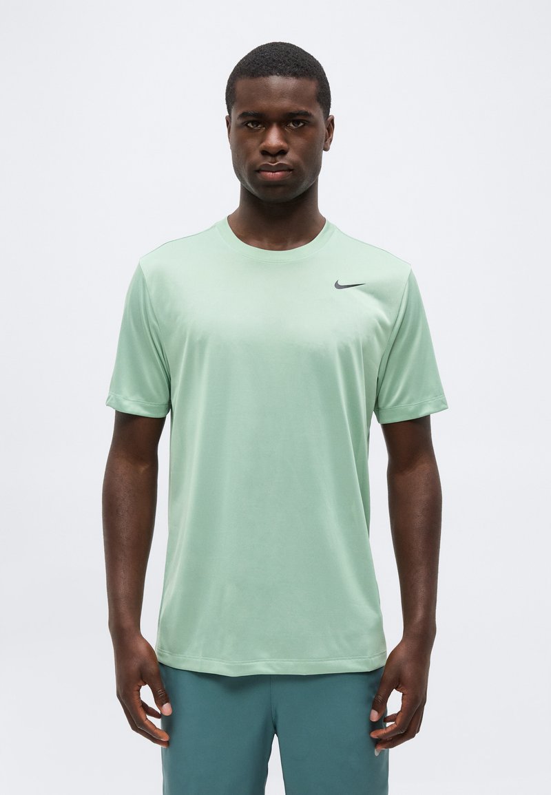 Νέος άνδρας με σκούρο δέρμα που φοράει ανοιχτοπράσινο αθλητικό μπλουζάκι Nike και μπλε-πράσινα σορτς, στέκεται μπροστά σε απλό λευκό φόντο.