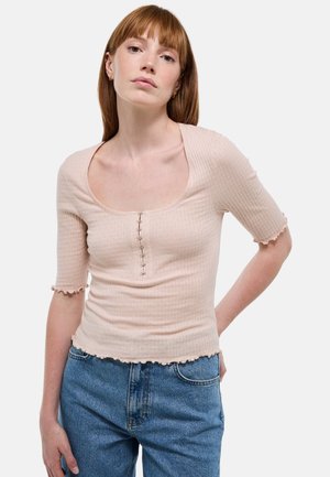 Haut côtelé rose pâle avec un large décolleté en scoop, des manches courtes et des bords volantés. Présente des boutons décoratifs sur le devant.