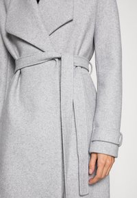 Cappotto di lana grigio con cintura, caratterizzato da un colletto a incavo, texture morbida e polsini arrotolati, che mette in risalto un design elegante e moderno.