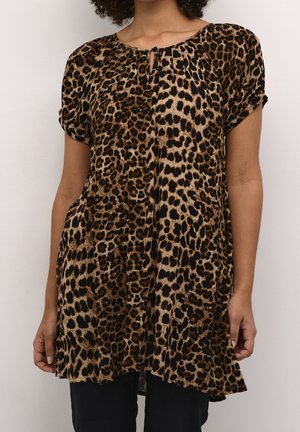 Vrouw draagt een korte mouw tuniek met luipaardprint en een keyhole-halslijn, gecombineerd met een zwarte broek tegen een effen lichte achtergrond.