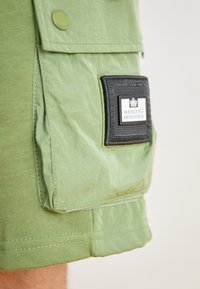 Grüne Cargo-Shorts mit strukturierter Oberfläche, ausgestattet mit einer Seitentasche und einem schwarzen Patch, auf dem "WEEKEND OFFENDER" steht. Die Nähte sind am Saum sichtbar.