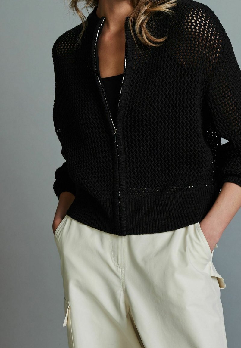Maglione nero con zip, caratterizzato da un design a maglia testurizzata, con maniche lunghe e polsini a costine, indossato con pantaloni beige chiaro.