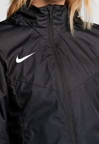 Zwarte Nike-jack met ritssluiting, hoge kraag en trekkoorden. Het materiaal is glad met een subtiele glans en een wit logo op de borst.