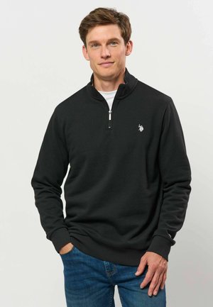 Homme portant un pull noir zippé sur une chemise blanche et un jean bleu, debout avec une main dans la poche sur un fond uni.