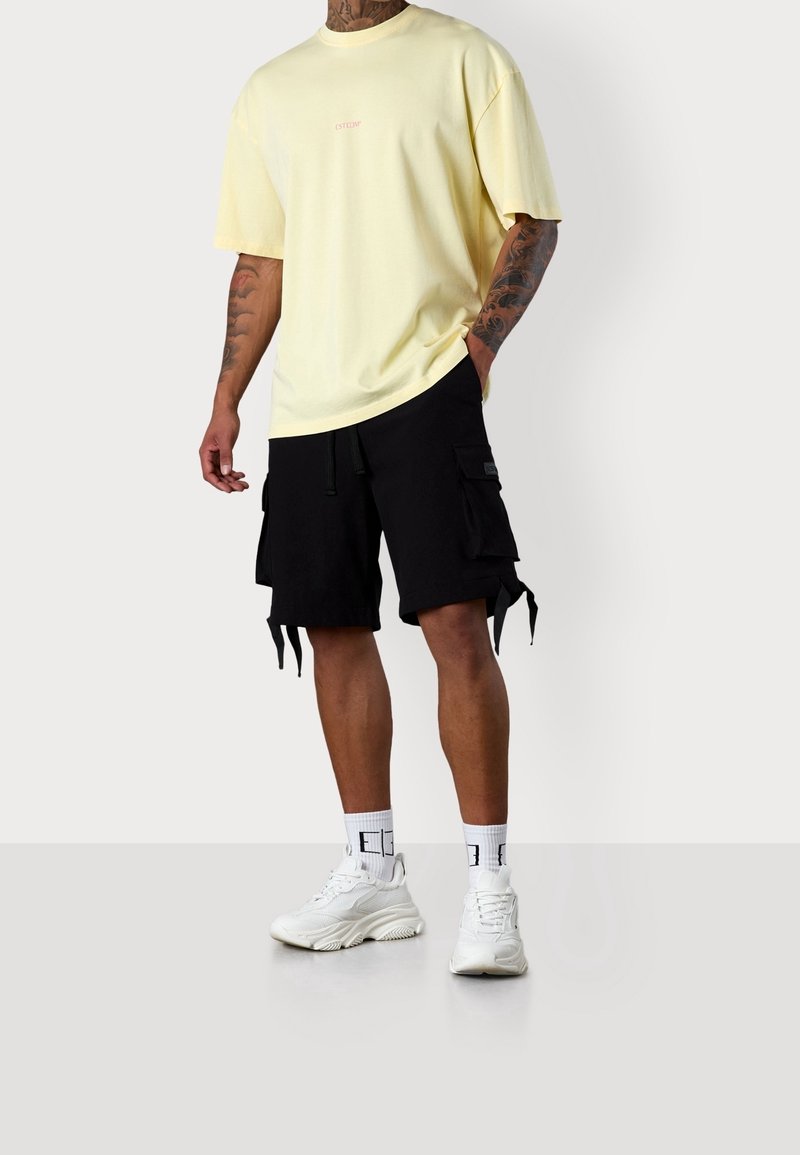 Gelbes übergroßes T-Shirt mit Logo, kombiniert mit schwarzen Cargo-Shorts mit Taschen und Schnüren. Weiße Sneakers und Socken mit grafischem Design.