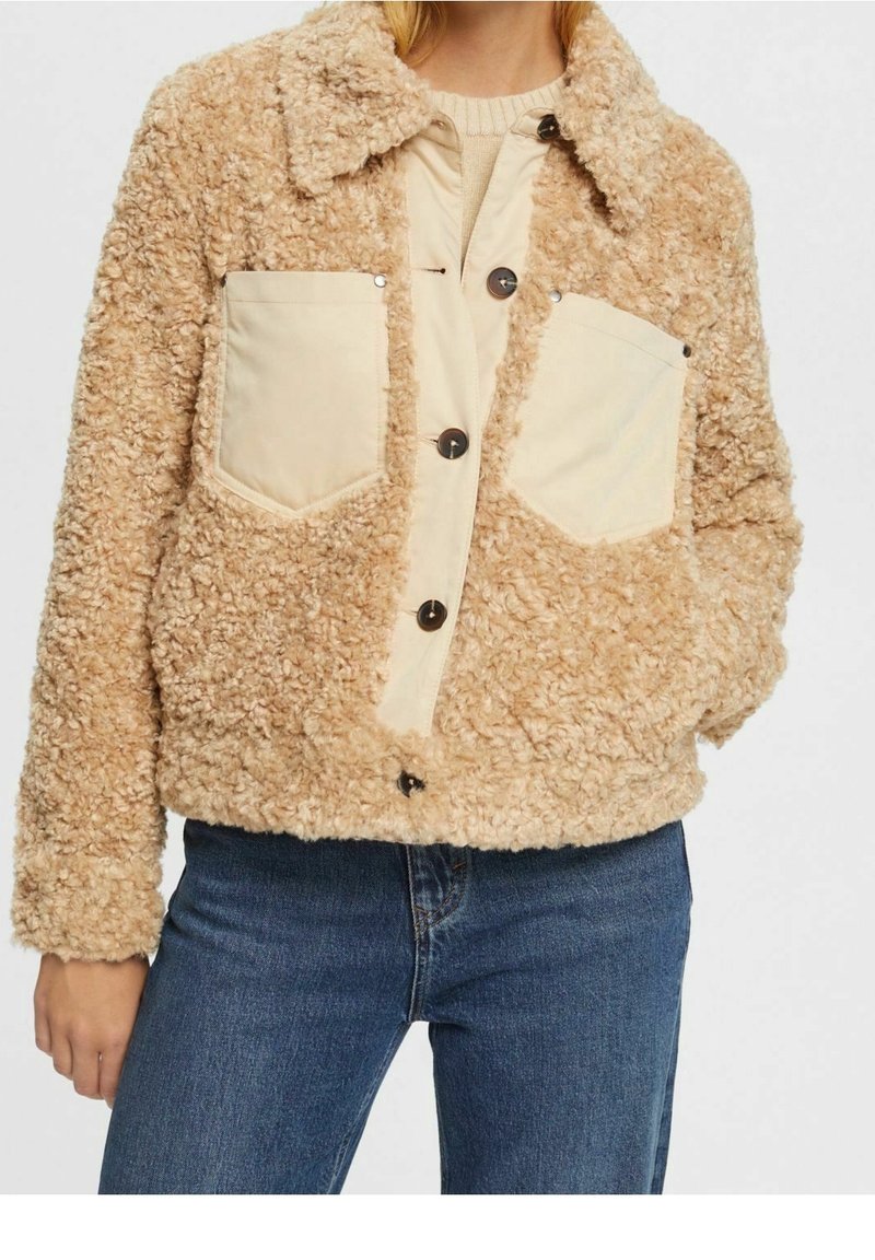 Veste en polaire marron et duveteuse avec un devant beige à boutons, deux poches poitrine et un col, portée avec un jean bleu.