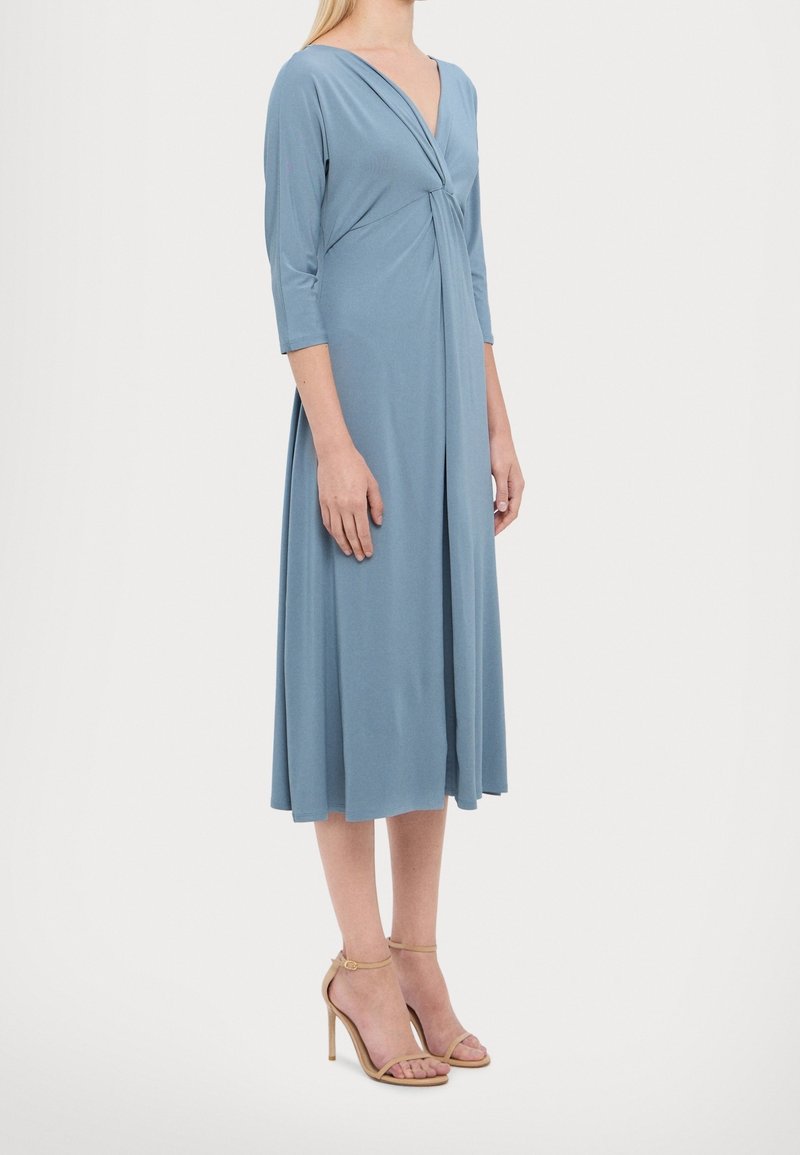 Femme portant une robe midi bleu clair avec manches 3/4, un décolleté profond en V et des sandales beige à talons hauts avec bride à la cheville, debout de profil.