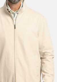 Chaqueta ligera beige con cuello alzado, cremallera frontal y dos bolsillos laterales. Tejido suave con un diseño minimalista.