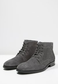 Pair of gray suede lace-up ankle boots with perforated detailing on the toe and black rubber soles on a white surface.