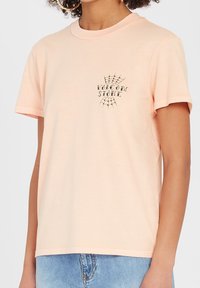 Korte mouwen perzikkleurig t-shirt van katoen, met een klein grafisch ontwerp van een spinnenweb en de tekst "Volcom Stone" op de voorkant.