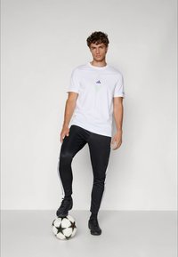 White cotton t-shirt with a small green and purple logo, paired with black athletic pants featuring white side stripes; black soccer ball at feet.