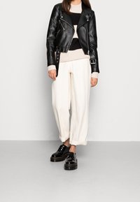 Schwarze, kurze Bikerjacke aus Leder mit silbernen Reißverschlüssen, cremefarbener Strickpullover, lockere, cremeweiße Hosen und schwarze Plateau-Schuhe.