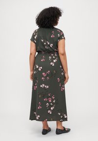 Maxi dress in dark green fabric with short sleeves, floral pattern in pink and brown, elastic waist, and worn with black slip-on shoes.