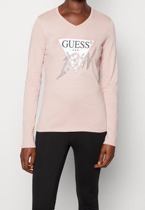 Personne portant un haut à manches longues rose clair avec un col en V, un logo Guess pailleté et un pantalon noir, debout devant un fond uni.